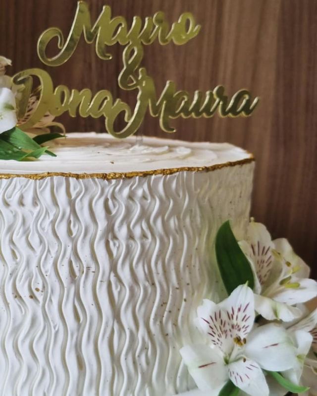 Topo dourado com os nomes Mayro e Dona Maura sobre bolo branco com flores